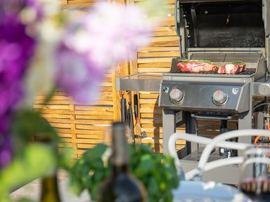 A barbecue grill with meat on the grill in an outdoor area with wooden fencing and a table with wine bottles in the foreground at Newquay Tamarisk Lodge on the Gannel in Newquay