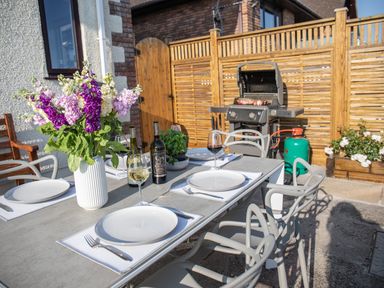 An outdoor dining table set with plates glasses and wine bottles next to a barbecue grill and wooden fence at Newquay Tamarisk Lodge on the Gannel in Newquay