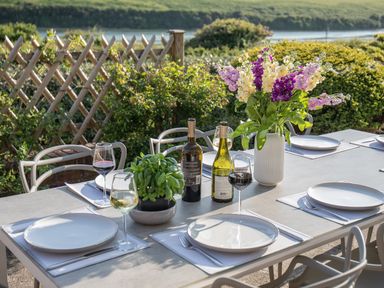 An outdoor dining table set with white plates wine glasses cutlery bottles of wine a vase with purple and yellow flowers and a potted green plant at Newquay Tamarisk Lodge on the Gannel in Newquay