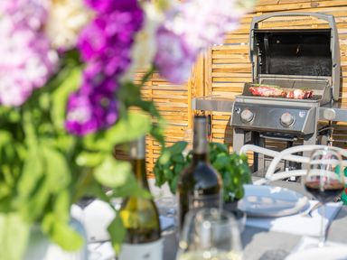 An outdoor dining setup with a wine bottle glass and flowers on a table and a grill with meat cooking in the background at Newquay Tamarisk Lodge on the Gannel in Newquay