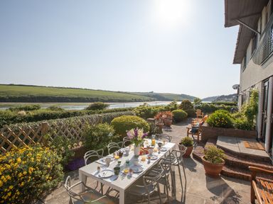 An outdoor patio with a dining table set for breakfast and chairs overlooking a river and green hills at Newquay Tamarisk Lodge on the Gannel in Newquay