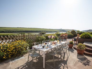 An outdoor patio dining area with a table set for breakfast and chairs overlooking a river and green fields at Newquay Tamarisk Lodge on the Gannel in Newquay