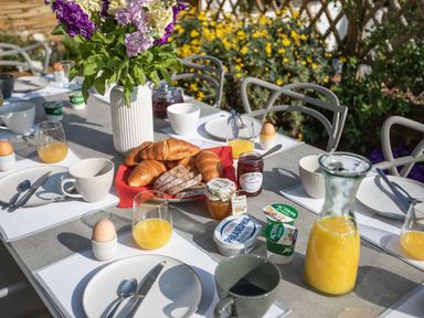An outdoor breakfast table set with plates cups glasses of orange juice soft boiled eggs croissants bread jars of jam yogurt and a vase of flowers at Newquay Tamarisk Lodge on the Gannel in Newquay