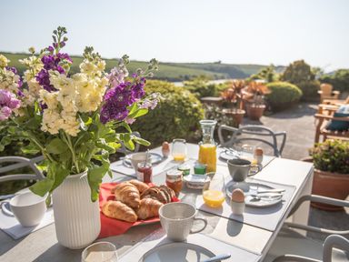 A patio table set with croissants juice eggs and cups with flowers in a vase outdoors at Newquay Tamarisk Lodge on the Gannel in Newquay