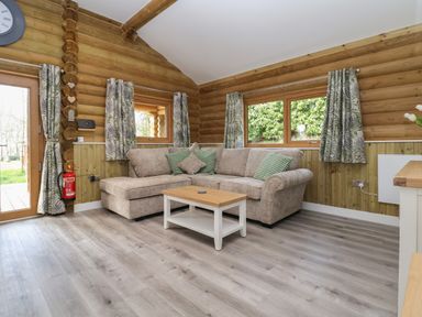 A living room with a beige sectional sofa green patterned cushions a wooden coffee table wood-paneled walls and windows with leafy curtains at Redwood Lodge in Ellesmere