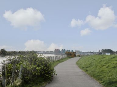 A paved pathway beside a river with grassy banks and bushes near the water at Cosy Cottage in Amble