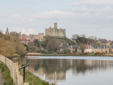 A river with houses and a castle on a hill in the background at Cosy Cottage in Amble