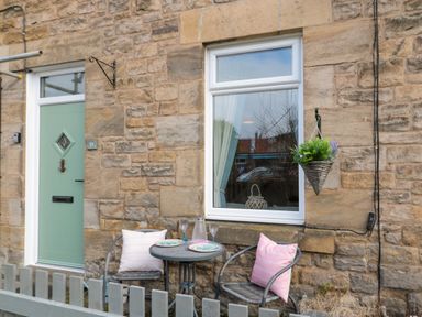 A small outdoor seating area with two chairs and a round table set against a stone wall with a green door and a window with a hanging plant at Cosy Cottage in Amble