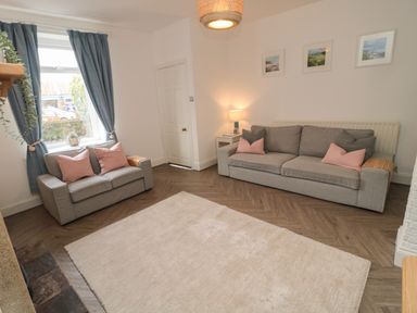 A living room with two gray sofas pink cushions a white rug wooden floor and blue curtains at Cosy Cottage in Amble