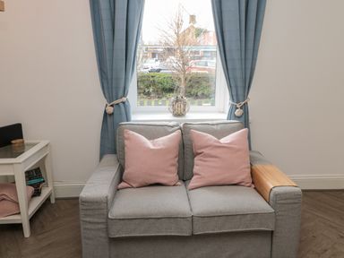 A grey sofa with two pink pillows in front of a window with blue curtains and a small white side table with books and a pink throw at Cosy Cottage in Amble