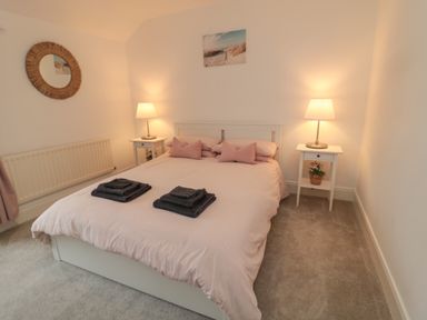 A bedroom with a double bed with pink pillows and blankets two bedside tables with lamps and flowers a circular mirror on the wall and a radiator at Cosy Cottage in Amble