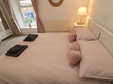 A bedroom with a double bed with pink bedding towels on the bed a bedside table with a lamp and a window with curtains and a flower vase at Cosy Cottage in Amble