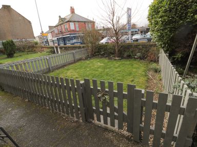 A small garden with a gray wooden fence and gate near a street with buildings and cars at Cosy Cottage in Amble