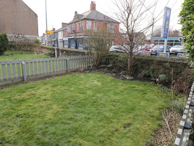 A small fenced garden with grass and shrubs next to a street with buildings and parked cars at Cosy Cottage in Amble