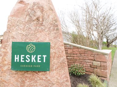 A green sign on a large rock that reads Hesket Caravan Park near a stone wall and plants at Rosie's Zen Den in High Hesket near Armathwaite