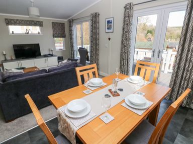 A dining area with a wooden table set for four with white dishes and wine glasses next to a living room with a dark sofa and a TV on a cabinet at Lodge 53 Stepaside near Kilgetty