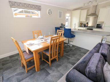 A dining area with a wooden table and four chairs next to a kitchen island with two blue bar stools and a gray sofa at Lodge 53 Stepaside near Kilgetty