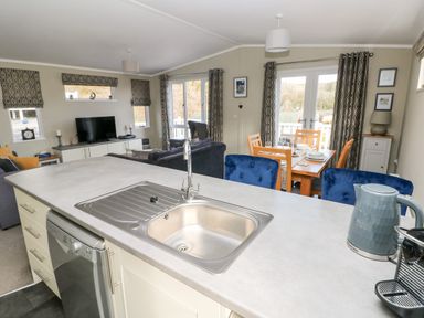 A kitchen island with a sink and kettle overlooking a living room with a television and a dining area with a wooden table and blue chairs at Lodge 53 Stepaside near Kilgetty