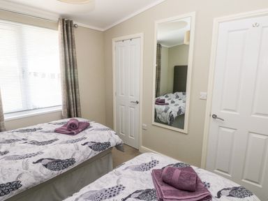 A bedroom with two single beds with peacock patterned bedding and folded towels in front of a window with curtains and blinds at Lodge 53 Stepaside near Kilgetty