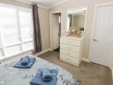 A bedroom with a bed with folded towels by a window with curtains and a chest of drawers with a mirror at Lodge 53 Stepaside near Kilgetty