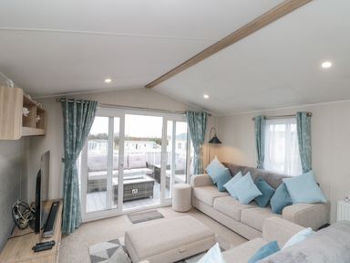 A living room with a beige sofa and ottoman with blue cushions and teal curtains by sliding glass doors opening to an outdoor seating area at Rowan Court 2 in Cayton