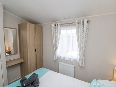 A bedroom with a bed a wooden wardrobe a wall-mounted mirror with a small shelf and a window with curtains at Rowan Court 2 in Cayton