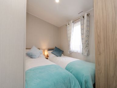 A bedroom with two single beds with blue blankets and pillows a lamp on a nightstand and a window with patterned curtains at Rowan Court 2 in Cayton