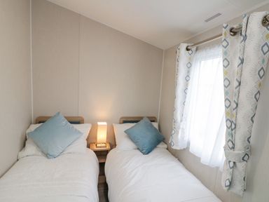 A bedroom with two single beds with blue pillows a bedside table lamp and a window with patterned curtains at Rowan Court 2 in Cayton