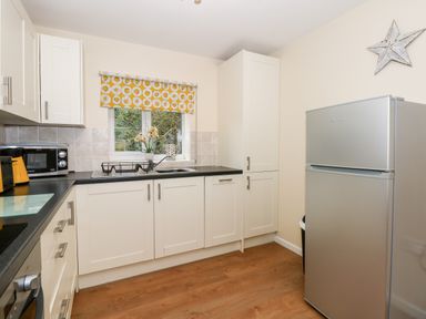 A kitchen with white cabinets black countertops a silver refrigerator and a window with a yellow patterned blind at 1 The Dell in Mundesley