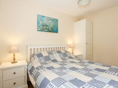 A bedroom with a bed with a blue and white checkered quilt two bedside tables with lamps a wardrobe and a painting on the wall at 1 The Dell in Mundesley