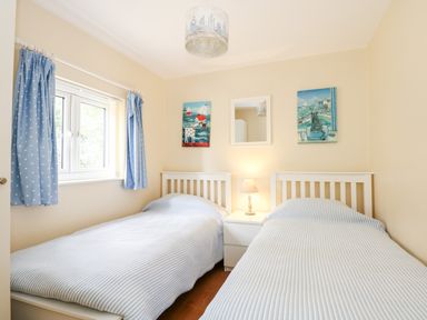 A bedroom with two single beds blue striped bedding a window with blue curtains and a white nightstand with a lamp at 1 The Dell in Mundesley