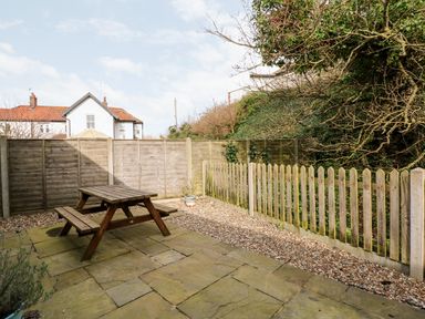 An outdoor patio with a wooden picnic table surrounded by a wooden fence and gravel at 1 The Dell in Mundesley