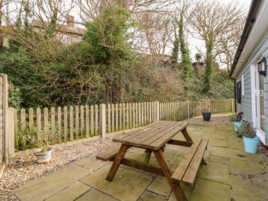 A patio with a wooden picnic table and potted plants next to a light blue house at 1 The Dell in Mundesley