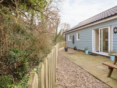A gravel and paved patio area beside a light blue house with a wooden fence and trees at 1 The Dell in Mundesley