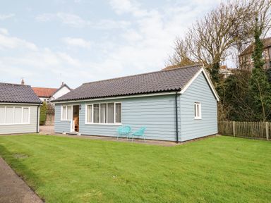A single-story house with blue siding and a tiled roof with two turquoise chairs on a patio and a lawn at 1 The Dell in Mundesley