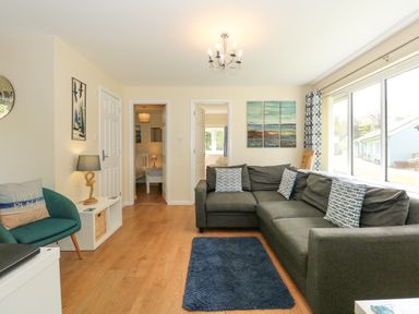 A living room with a grey sectional sofa and patterned cushions next to large windows at 1 The Dell in Mundesley