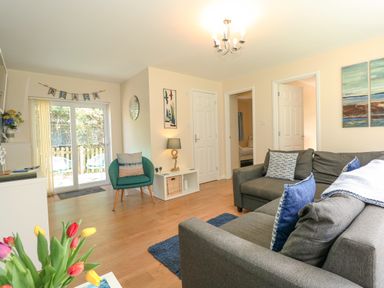 A living room with a grey sofa, blue cushions, a green armchair, sliding glass doors, and a white cabinet at 1 The Dell in Mundesley