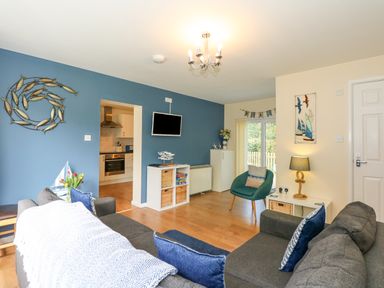 A living room with blue and beige walls a gray sofa with blue pillows a green chair a wall mounted TV and an open kitchen door at 1 The Dell in Mundesley