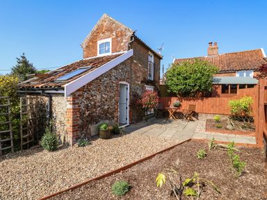 A garden area with gravel and paved patio with table and chairs next to a brick and flint cottage at Admiral Cottage in Blaxhall near Snape Suffolk