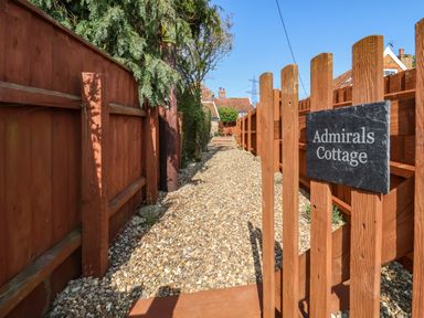 A gravel pathway with wooden fences on both sides and a wooden gate with a sign that reads Admirals Cottage at Admiral Cottage in Blaxhall near Snape Suffolk