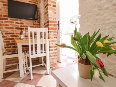 A dining area with white chairs and a wooden table set against a brick wall with a mounted TV and a vase of tulips on a white surface at Admiral Cottage in Blaxhall near Snape Suffolk