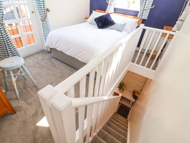 A bedroom with a bed pillows stool window with curtains and stair railing at Admiral Cottage in Blaxhall near Snape Suffolk