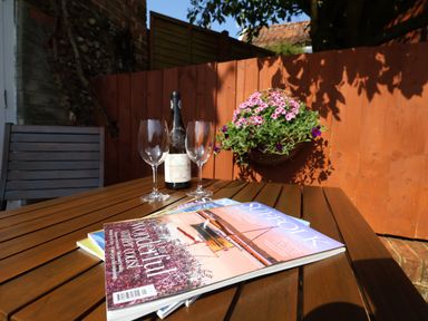 A wooden outdoor table with a bottle of wine two wine glasses magazines and a hanging basket of flowers at Admiral Cottage in Blaxhall near Snape Suffolk