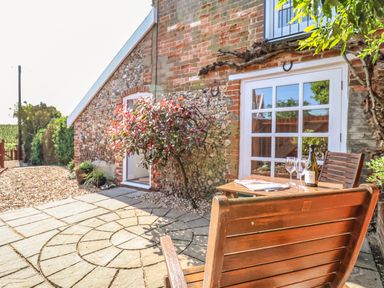 An outdoor patio with wooden chairs and table set near a stone and brick house wall with a glass door at Admiral Cottage in Blaxhall near Snape Suffolk