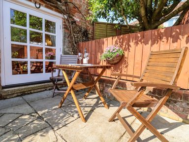 An outdoor patio area with two wooden chairs a wooden table a bottle of wine and two glasses on the table and a flower basket hanging on a wooden fence at Admiral Cottage in Blaxhall near Snape Suffolk