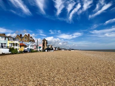 A pebble beach with buildings along the shore and a blue sky with clouds at Admiral Cottage in Blaxhall near Snape Suffolk