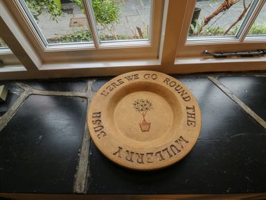 A ceramic plate with a potted plant illustration and the words here we go round the mulberry bush on a black stone windowsill at Mulberry Tree Appledore