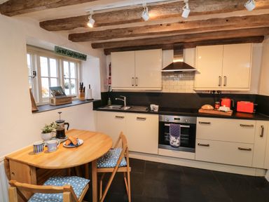 A kitchen with cream cabinets wooden beams on the ceiling a wooden table with two chairs and blue cushions at Pear Tree Cottage in Appledore