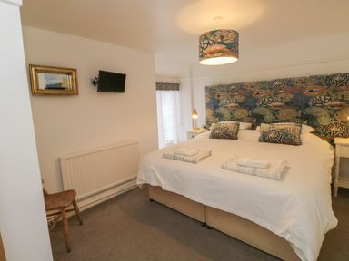 A bedroom with a large bed with patterned headboard and cushions two bedside tables with lamps a small wall mounted tv a wooden chair and a window at Pear Tree Cottage in Appledore