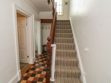 A hallway with checkered tile floor and a striped carpeted staircase with wooden banister at Lands House in Appledore